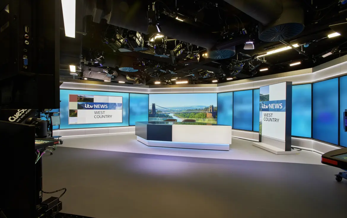A modern television news studio set with a sleek, professional design. In the centre, a curved news desk with a white and dark grey base features an integrated screen displaying a landscape image. Behind the desk, a wide wall of large blue-toned display panels wraps around the studio, creating a bright, high-tech backdrop.  On the right side, a vertical freestanding screen shows the text “itv NEWS WEST COUNTRY” in bold lettering. A larger horizontal screen on the left wall repeats the same branding. The studio floor is smooth and grey, with subtle lighting creating soft reflections.  Above, the ceiling is open, revealing an array of lighting rigs, cameras, cables and ventilation systems, all arranged in a dense technical grid. Long linear light fixtures run parallel across the ceiling, illuminating the set evenly. On the far left edge of the image, part of a large black camera or broadcast equipment rig is visible, reinforcing the production environment. The overall space feels polished, controlled and designed for live broadcasting.