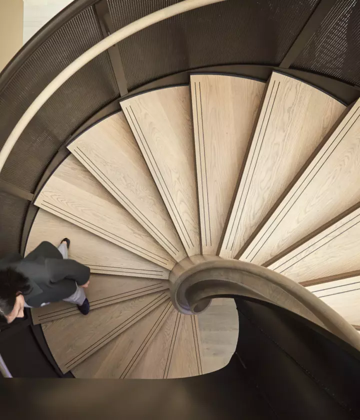 Interior view featuring a spiral staircase with wooden steps and a dark metal railing. The staircase is a central architectural element to a modern and stylish office. The design emphasises natural materials and curved forms, contributing to a contemporary and visually engaging environment.