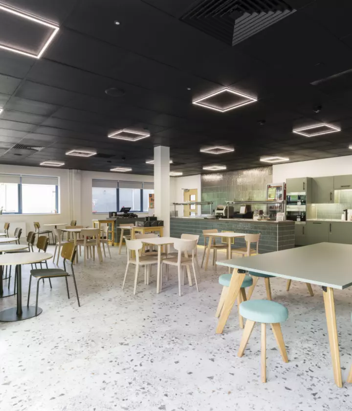 A bright, modern office cafeteria featuring mixed seating options including tables with chairs, a long wall bench and a communal table with stools. Square ceiling lights and large windows provide ample illumination. At the back, a counter area houses coffee machines and kitchen equipment, blending functionality with a welcoming atmosphere.