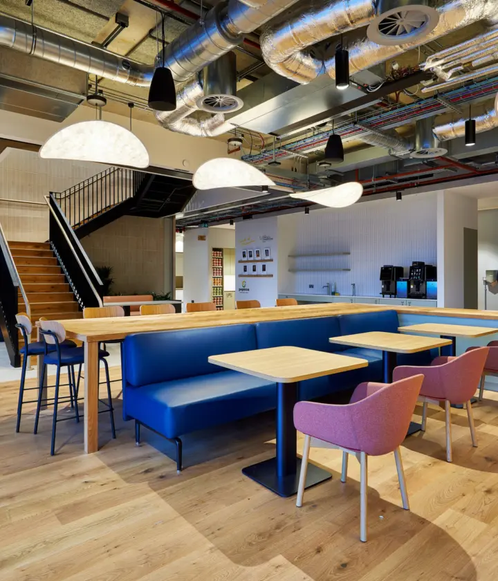 Open plan office breakout area with long communal tables, blue upholstered bench seating and pink chairs on a light wood floor. Exposed ductwork and cables run across the ceiling with large pendant lights. A staircase with black railings leads to an upper level, while a kitchenette with coffee machines sits to the rear.