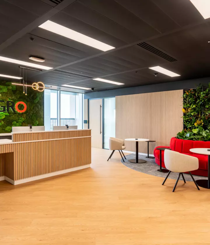 A modern office reception area with wooden flooring and ceiling. On the left, a wooden reception desk with white accents sits in front of a green wall displaying the text "SEGRO". On the right, a seating area includes red cushioned seats around small round tables with beige chairs. Another green wall with plants is positioned behind the seating area. Large windows allow natural light to enter the space.