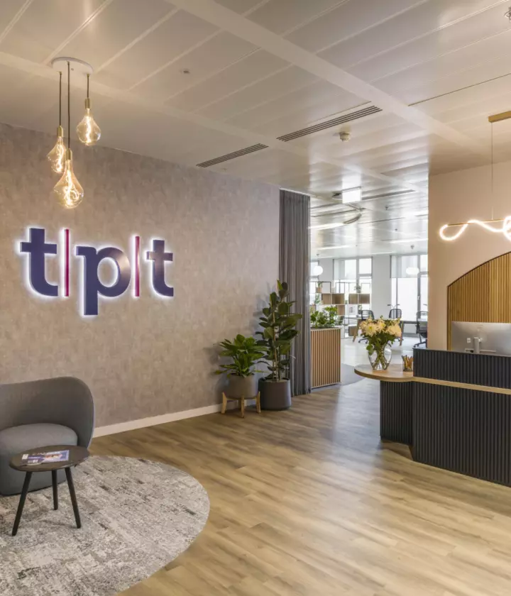 Modern office reception area with wooden flooring, bespoke reception desk, illuminated company logo, grey armchairs on a circular rug, pendant lighting and open plan workspace in the background