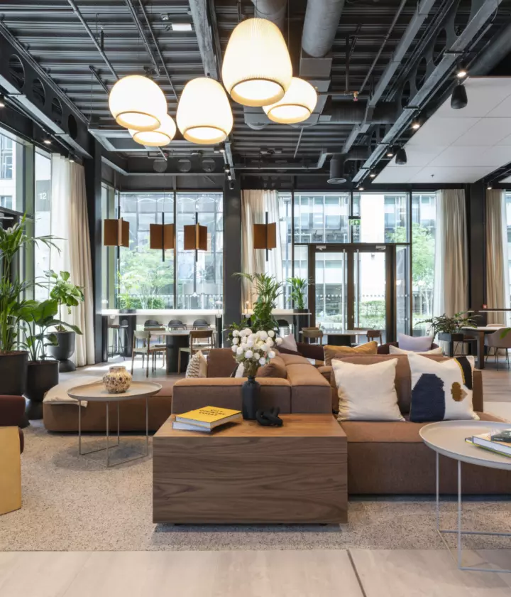 Modern office lounge area featuring a mix of contemporary furniture, including modular sofas, armchairs, and round coffee tables arranged on a textured rug. The space has large floor-to-ceiling windows for natural light, complemented by pendant lighting and exposed ceiling elements for an industrial aesthetic. Indoor plants add greenery, while neutral tones with wood accents create a warm, inviting atmosphere suitable for collaborative work and informal meetings