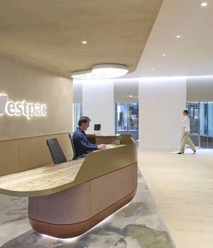 A contemporary office reception area created for Westpac, featuring a curved desk with a stone top and soft timber finishes. A receptionist is seated at the desk while a person walks through the bright open space beyond. Built-in storage, natural planting and subtle lighting contribute to a calm workplace environment that reflects modern office fit out and office design principles.