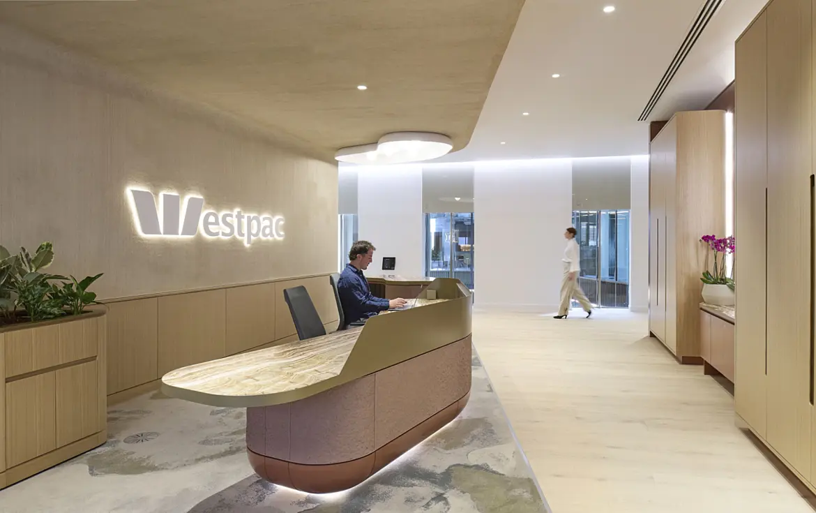 A contemporary office reception area created for Westpac, featuring a curved desk with a stone top and soft timber finishes. A receptionist is seated at the desk while a person walks through the bright open space beyond. Built-in storage, natural planting and subtle lighting contribute to a calm workplace environment that reflects modern office fit out and office design principles.