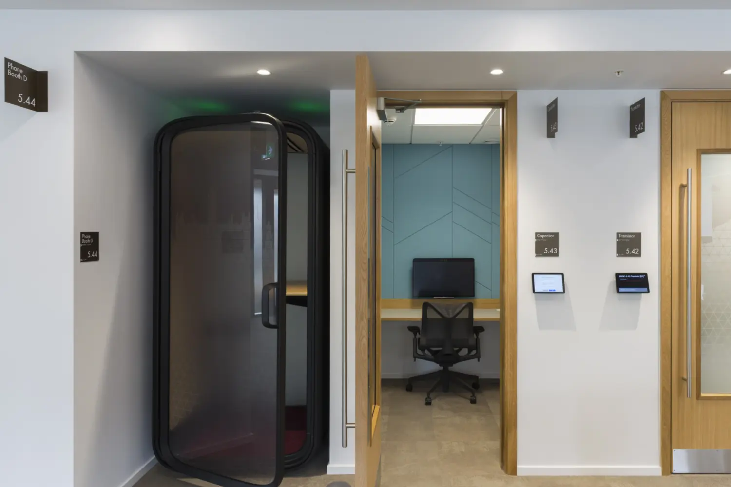 A quiet office corridor with light flooring and recessed ceiling lights. On the left is a small enclosed phone booth with a curved glass door. Next to it is an open doorway leading to a small workspace with a desk, a monitor and a task chair. Room signs and booking screens are mounted on the wall beside the doors.