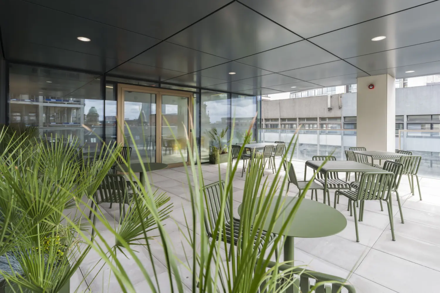 A covered outdoor terrace with tiled flooring and a dark ceiling. Green metal tables and chairs are spaced across the area. Potted plants with tall leaves sit in the foreground. Glass doors lead back inside the building and the terrace overlooks nearby rooftops and concrete structures.