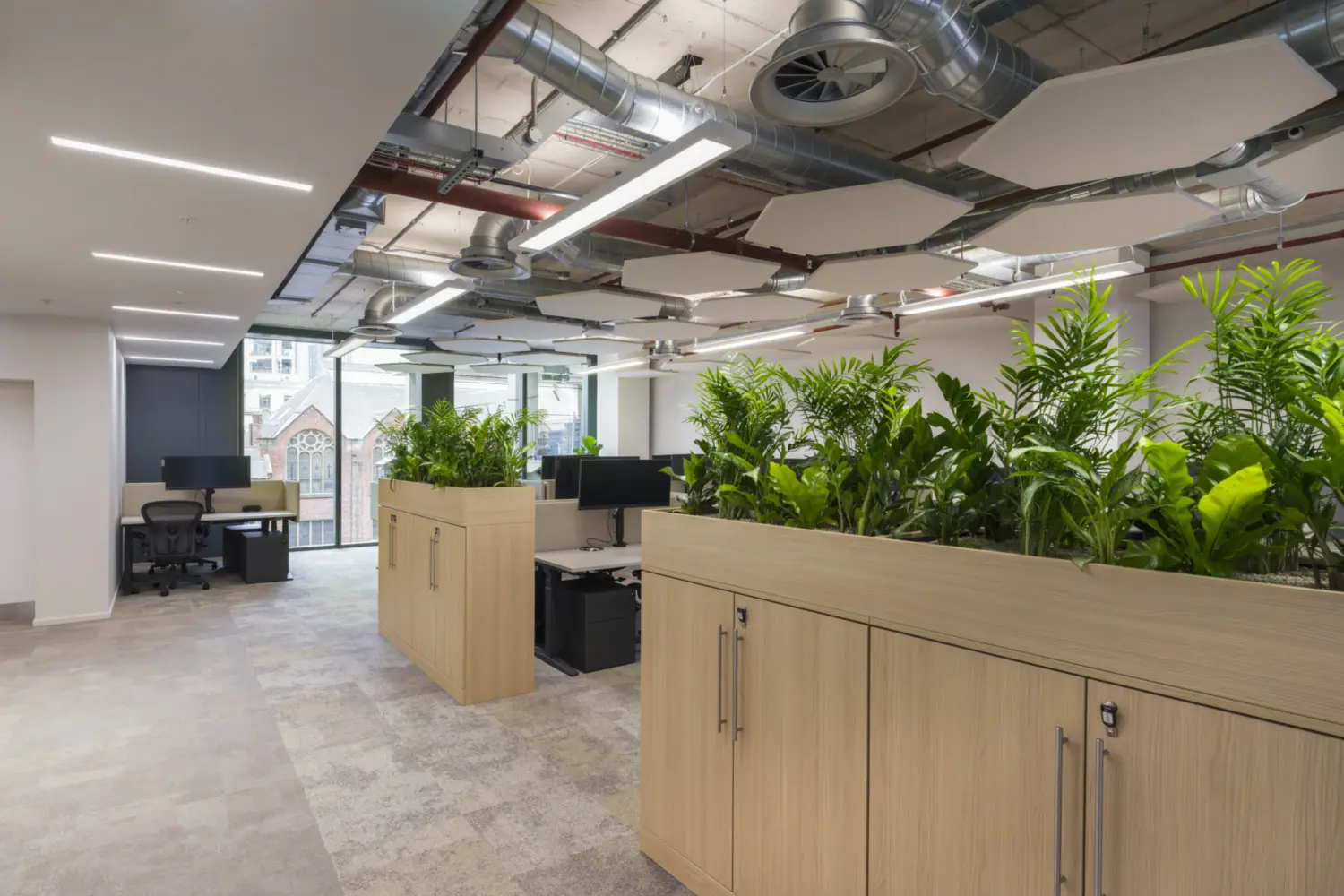 An open plan office with light flooring and exposed services in the ceiling. Rows of low storage units topped with dense green plants divide the workspace. Behind them are desks with monitors and task chairs. Large windows on the far side bring in natural light and offer a view of nearby buildings.