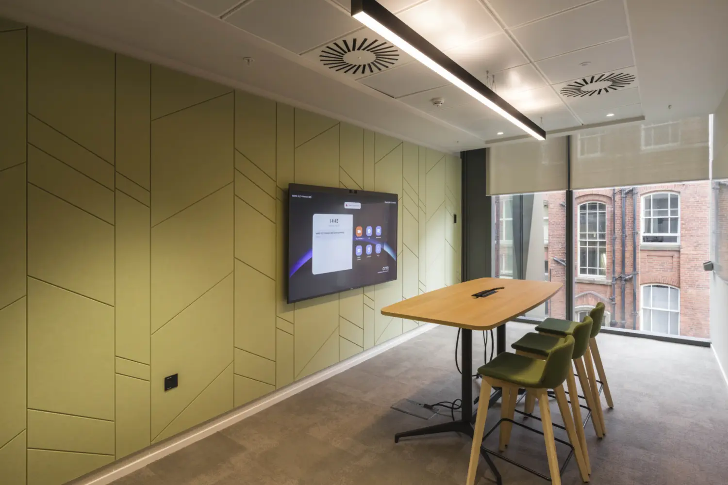 A small meeting room with pale green acoustic wall panels and a wall mounted screen displaying a video conferencing interface. A high table with four green stools sits in the centre of the room. Large windows on the right let in natural light and show the brick exterior of a nearby building. Ceiling lights and ventilation panels run above the space.