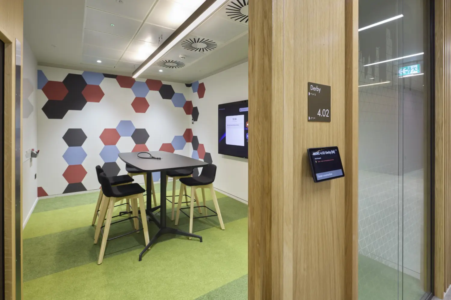 A small meeting room with a patterned feature wall made of red, blue and black hexagons. A high table with four stools sits on green carpet tiles. A wall mounted screen is positioned opposite the entrance. The room has a light ceiling with recessed lights and ventilation panels and the doorway features a wooden frame with a booking screen mounted outside.
