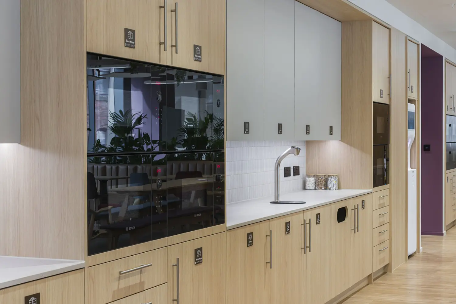 A modern office kitchen with light wood cabinets, integrated appliances and a long run of storage. White tiles form a small splashback above a counter with a tap and sink. The space has light wood flooring and recessed ceiling lights and reflections in the glossy cabinet doors show plants and seating in the adjoining area.