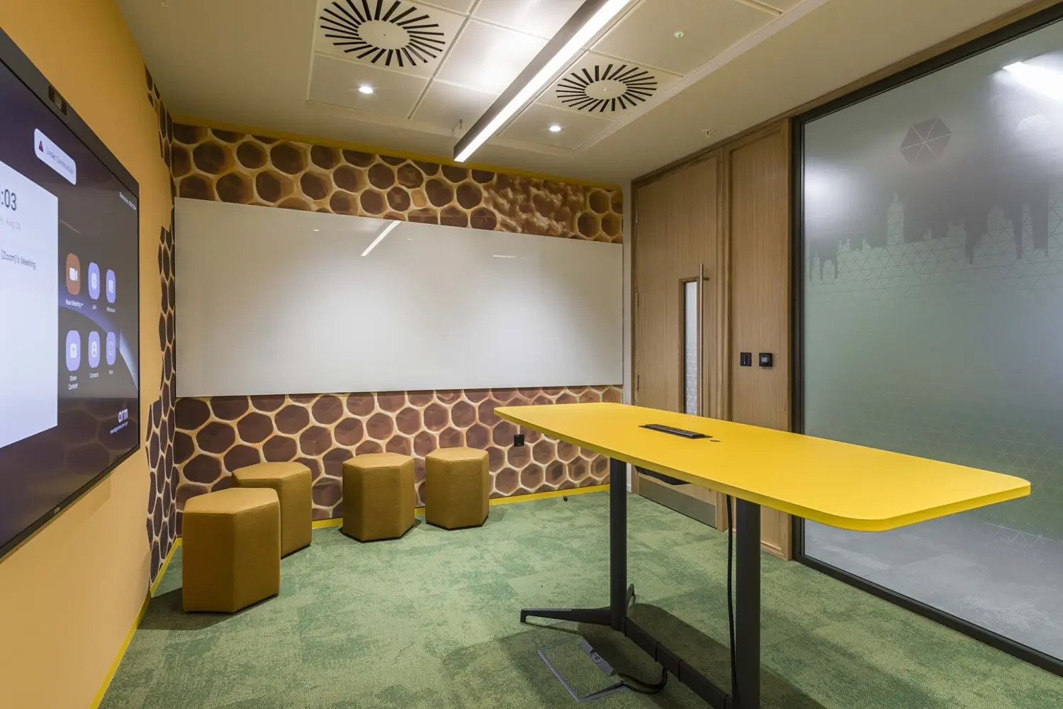 A small meeting room with honeycomb patterned wall panels and a large whiteboard. A bright yellow high table stands in the centre with four matching stools. A wall mounted screen sits opposite a frosted glass panel that features a subtle city skyline graphic. The room has green carpet tiles and ceiling lights with ventilation panels.