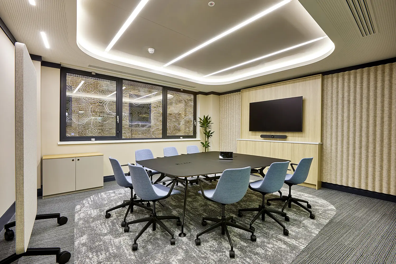 Modern meeting room featuring a rectangular black table surrounded by eight wheeled chairs upholstered in soft blue fabric. The room is finished with neutral tones and textured carpet flooring in grey, with light wood panelling and acoustic wall coverings adding warmth.  At the far end, a large wall-mounted display screen sits above a slim media unit, accompanied by a soundbar, with a tall indoor plant positioned in the corner. To the left, a row of windows fitted with decorative topographic pattern film allows filtered natural light into the space, with a low storage unit beneath.  The ceiling design features curved edges with recessed linear LED lighting forming clean geometric lines, alongside integrated ventilation and sensors. A freestanding acoustic screen is positioned in the foreground, contributing to privacy and sound control within the room.
