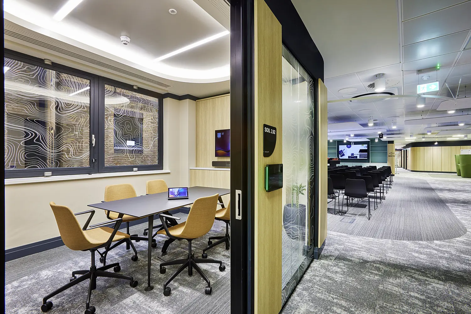 Modern office interior featuring a small meeting room in the foreground with a rectangular black table and four wheeled chairs upholstered in warm beige fabric. A tablet sits on the table, suggesting a collaborative or hybrid meeting setup. The room is enclosed by glass panels with a decorative topographic line pattern and framed in dark metal, with light wood wall finishes adding warmth.  Beyond the glass partition, an open area extends into a larger presentation or training space with rows of black chairs arranged facing a large screen. The ceiling throughout features curved, recessed lighting and integrated ventilation, while the flooring transitions between patterned carpet tiles in grey tones. A door panel with signage is visible on the partition, and natural light filters in through windows on the left, creating a bright and contemporary workspace.