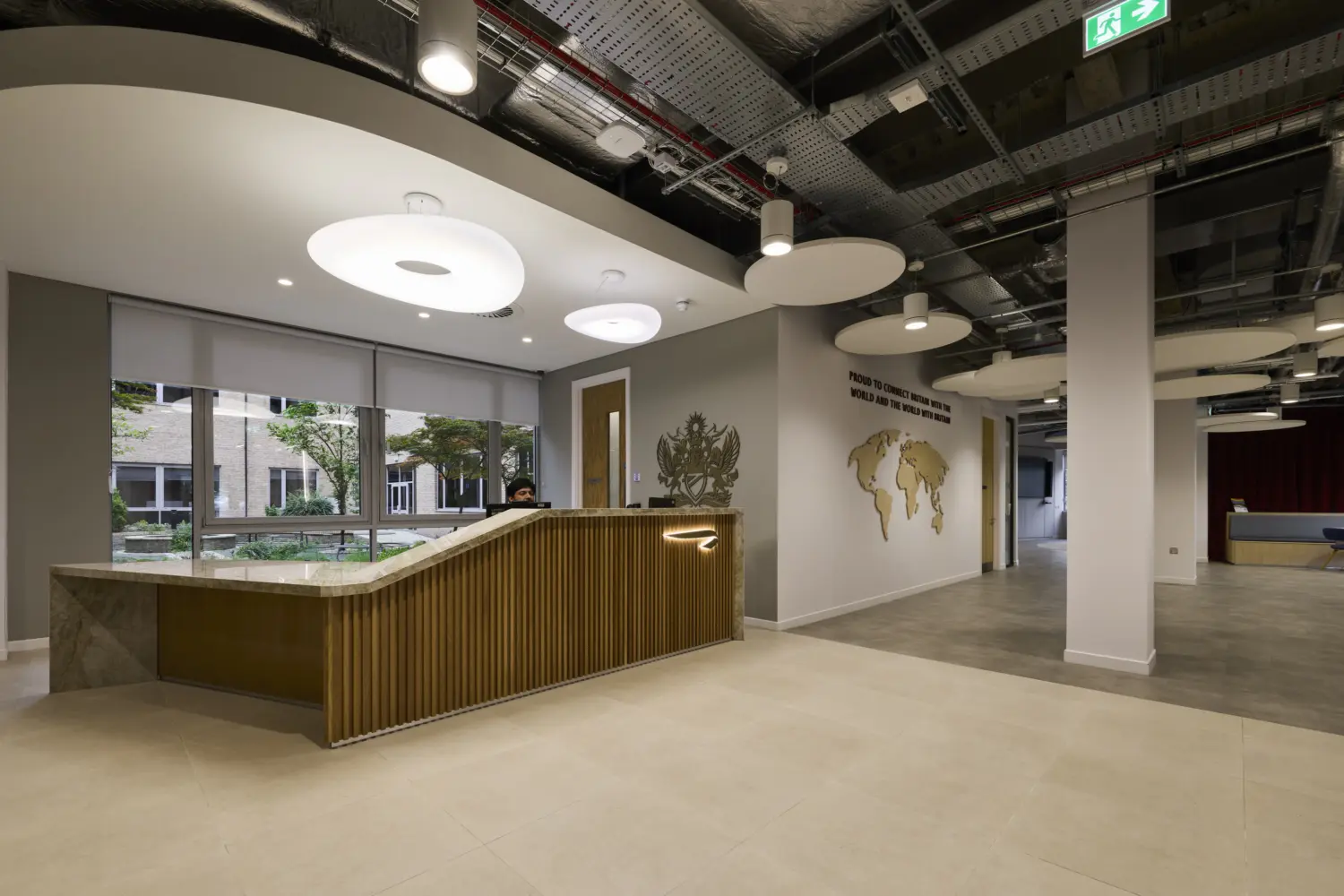 Modern office reception area featuring an angular timber-fronted welcome desk with a stone countertop, positioned beside large windows overlooking a landscaped courtyard. Circular acoustic ceiling panels and exposed building services create a contemporary aesthetic, while a timber world map feature wall and branded signage are visible in the background. The open-plan space connects to collaborative areas beyond, including a presentation zone with tiered seating and red curtains.