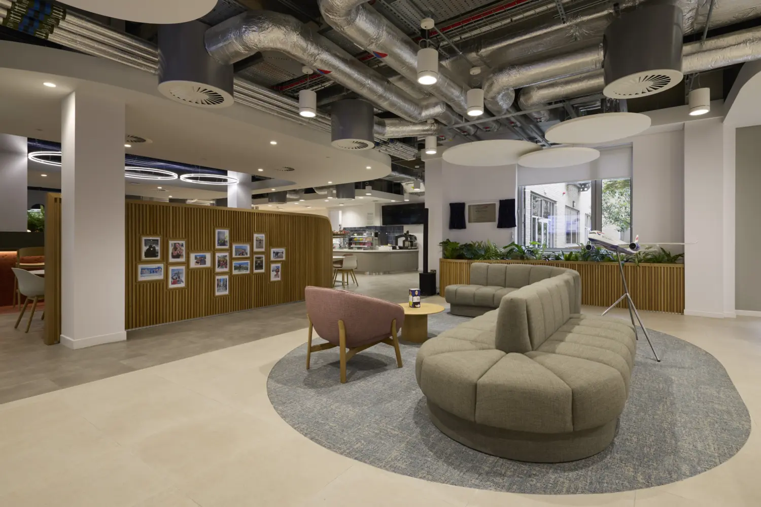Modern office breakout space with curved upholstered seating arranged around a circular rug, a lounge chair and coffee table in the foreground. A timber feature wall displaying framed photographs separates the area from a café and kitchen space in the background. Exposed services and acoustic ceiling panels create an industrial feel, while planting and large windows bring natural light into the interior.