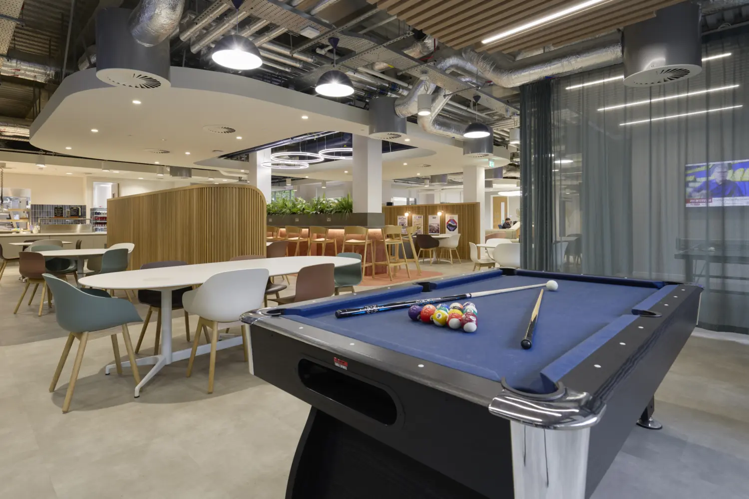 Modern office café and social space featuring a blue pool table in the foreground, surrounded by dining tables, lounge seating and high benches. Curved timber screening and integrated planting help define different zones within the open-plan area, while exposed services, pendant lighting and acoustic ceiling features create a contemporary industrial aesthetic. A large curtain divider separates part of the space, with kitchen and refreshment facilities visible in the background.