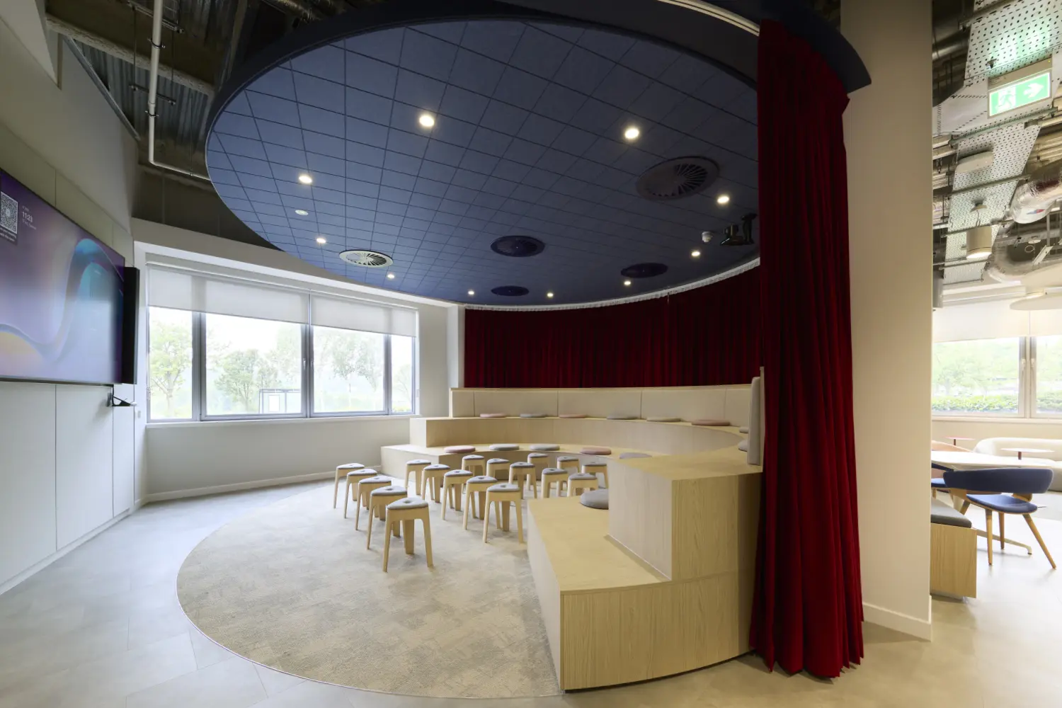 Semi-circular presentation town hall space within a modern office, featuring tiered timber seating, rows of stools and a large wall-mounted screen. Rich red curtains partially enclose the area, while a circular acoustic ceiling feature defines the space overhead. Large windows provide natural light, and adjacent lounge seating can be seen beyond the open edge of the room.