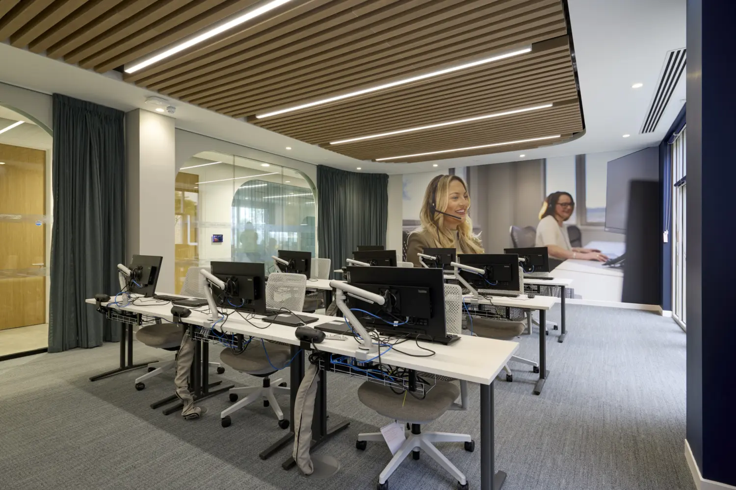 Modern office workspace with rows of sit-stand desks and computer workstations arranged in an open-plan setting. A feature wall graphic showing customer service staff spans the far end of the room, while glazed meeting rooms with arched detailing line one side. A timber slatted ceiling with integrated linear lighting adds warmth to the space, and full-height windows provide natural light along the perimeter.