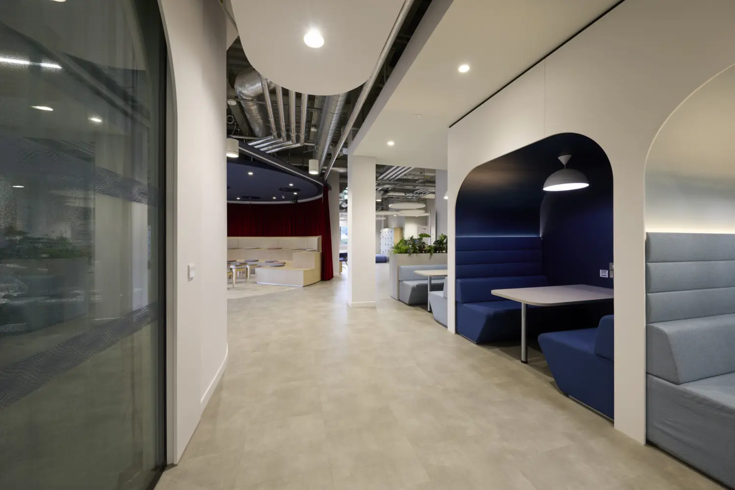 Modern office circulation space featuring a series of upholstered meeting booths with arched surrounds and integrated pendant lighting. The blue and grey booths provide informal areas for collaboration and focused work along a wide central walkway. In the background, a semi-circular presentation space with tiered seating and red curtains is visible, while exposed services, ceiling rafts and planting contribute to the contemporary workplace design.