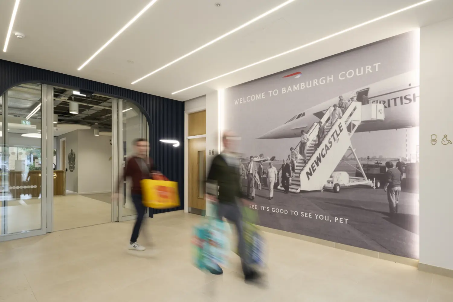 Modern office reception area featuring a large black and white heritage mural of passengers boarding a British Airways aircraft, with illuminated signage reading “Welcome to Bamburgh Court”. Arched glazed entrance doors framed by dark blue wall panelling lead into the workplace beyond. Two blurred figures walking through the space add a sense of movement, while recessed linear lighting and a neutral colour palette create a bright, contemporary arrival experience.