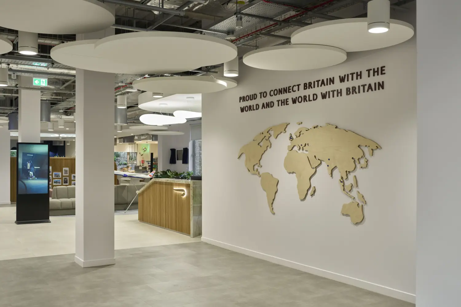 Modern office collaboration area featuring a large timber world map mounted on a white feature wall beneath the words, “Proud to connect Britain with the world and the world with Britain”. Circular acoustic ceiling panels and suspended lighting create visual interest overhead, while adjacent lounge seating, planting and timber detailing contribute to a welcoming, contemporary workplace environment. A digital display screen and open circulation space are visible in the foreground.