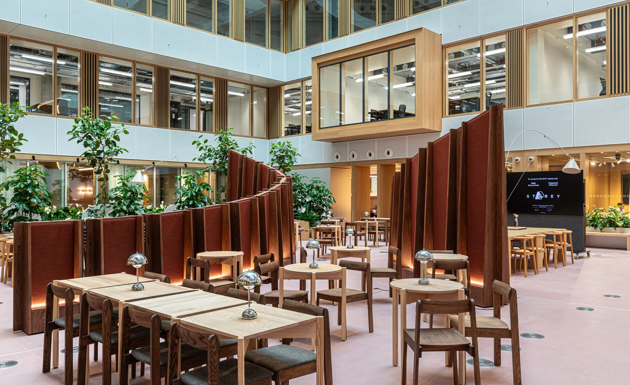 Modern open-plan office atrium with high ceilings and large windows. The space features wooden tables and chairs arranged in clusters, separated by curved, freestanding partitions in a reddish-brown fabric. Several green plants add a natural element throughout the area. A large digital screen is visible on the right wall, and an enclosed glass meeting room is suspended above the ground level. The overall design combines wood, glass and greenery for a contemporary and inviting atmosphere.
