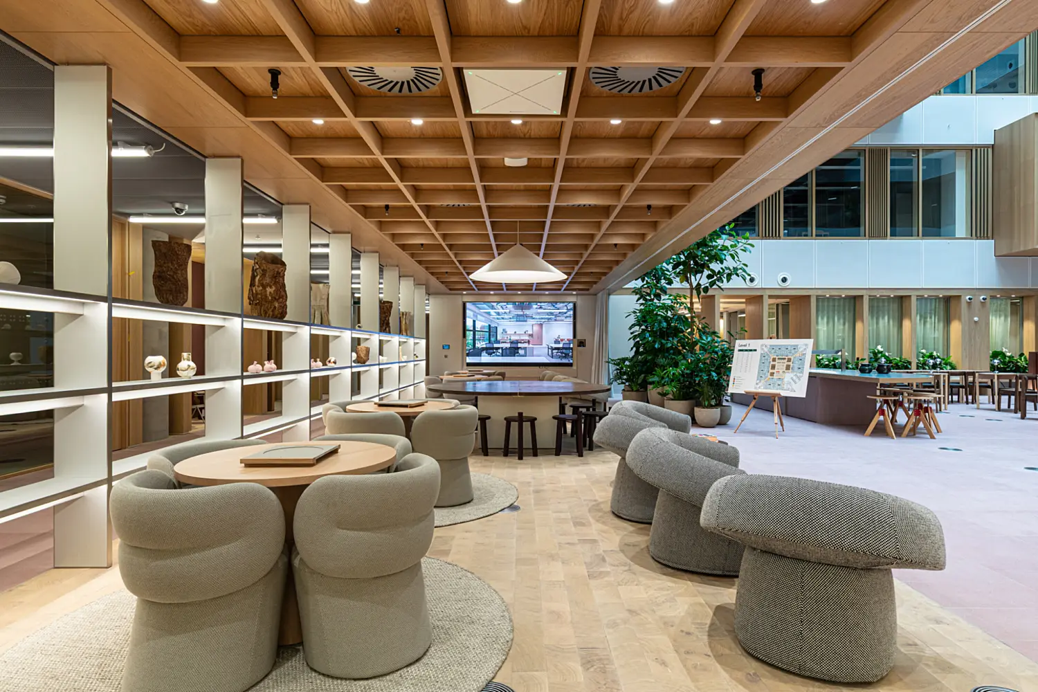 Warm and contemporary communal space within an office atrium featuring soft upholstered lounge chairs around circular tables. Timber shelving displays objects along one wall, while a coffered wooden ceiling adds texture overhead. The open area looks onto the atrium with planting and workspace visible beyond.