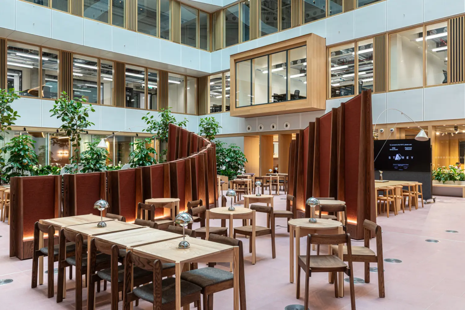 Modern open-plan office atrium with high ceilings and large windows. The space features wooden tables and chairs arranged in clusters, separated by curved, freestanding partitions in a reddish-brown fabric. Several green plants add a natural element throughout the area. A large digital screen is visible on the right wall, and an enclosed glass meeting room is suspended above the ground level. The overall design combines wood, glass and greenery for a contemporary and inviting atmosphere.