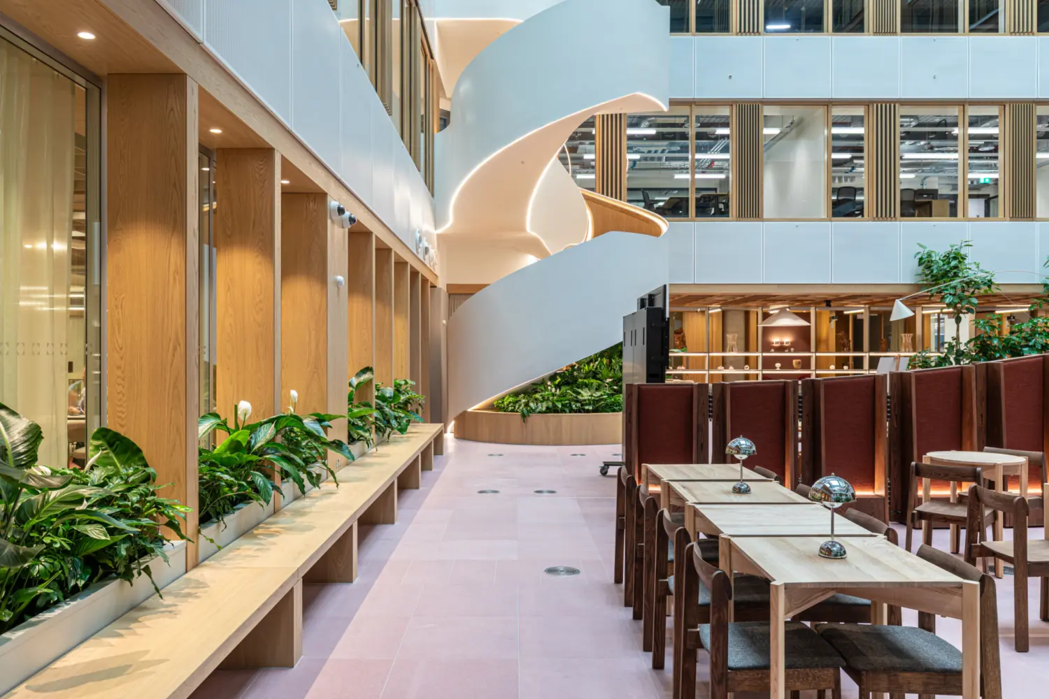 Modern office atrium interior featuring a sculptural white spiral staircase rising through the space. Wooden tables and chairs sit alongside curved partition screens, with integrated planting along a timber bench. Glazed offices line the upper floors, creating a bright and contemporary workplace setting
