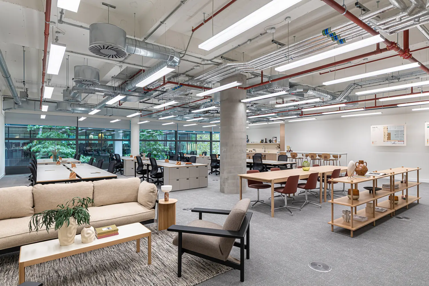 Open plan office workspace with exposed services and a high ceiling. The space includes bench desks with task chairs, a relaxed seating area with sofas and armchairs, and a long communal table. Large windows overlook greenery, bringing natural light into the contemporary working environment.