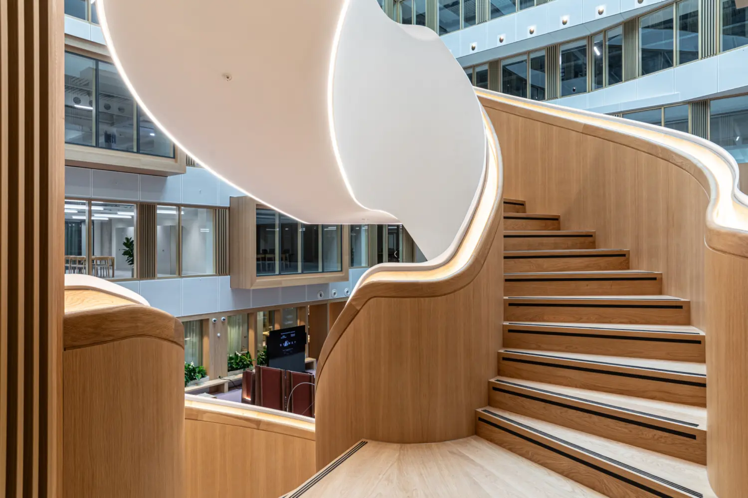 Close up view of a sculptural timber staircase within an office atrium, featuring curved wooden balustrades and integrated lighting. The staircase rises through the centre of a bright multi level space, with glazed offices and planting visible in the background.