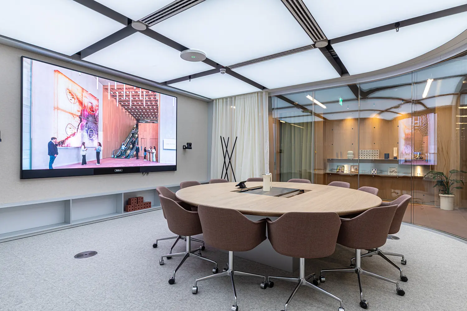 Modern meeting room with a round wooden table surrounded by upholstered task chairs. A large wall mounted screen displays digital content, while glazed partitions and sheer curtains separate the space from adjacent offices. Soft overhead lighting and neutral finishes create a calm and professional setting.