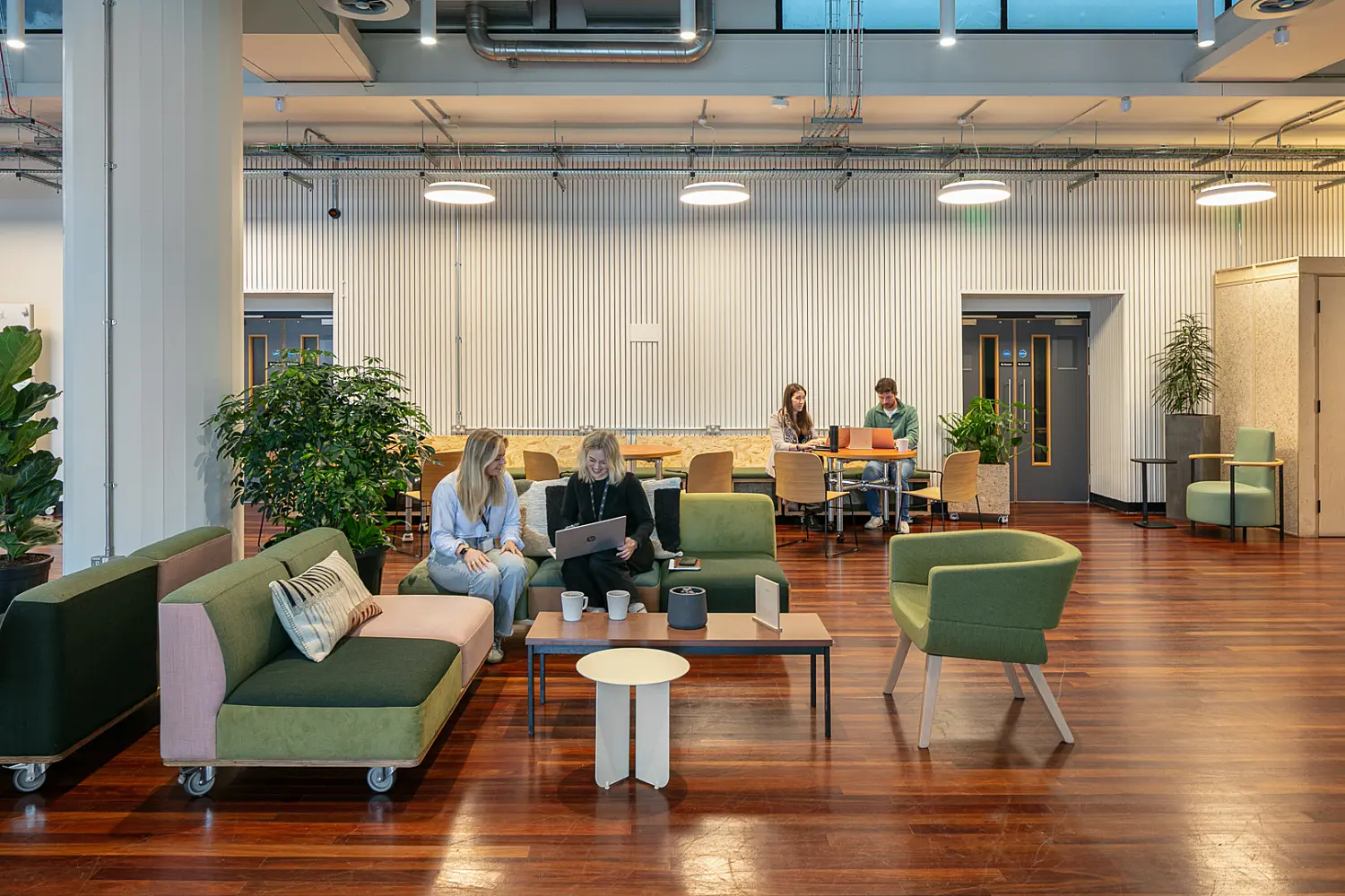 Modern workplace lounge area with polished wood flooring, green upholstered seating and coffee tables arranged for informal meetings. Two people are seated on a sofa working on a laptop, while two others sit at a table in the background. Large indoor plants, circular pendant lights and exposed services contribute to a contemporary industrial-style interior. The space includes breakout seating, collaborative work areas and light-coloured feature walls.