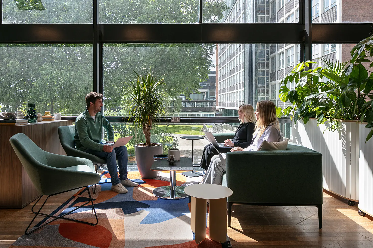Informal workplace collaboration area set beside floor-to-ceiling windows overlooking trees and neighbouring buildings. Three people are seated on lounge chairs and a sofa around small tables, working on laptops and holding a discussion. The space features green upholstered seating, a colourful geometric rug, polished wood flooring and large indoor plants positioned throughout. Natural daylight fills the area, creating a bright and comfortable setting for meetings, focused work and informal collaboration.