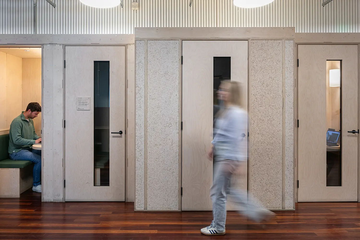 Row of timber-clad focus booths and meeting rooms within a contemporary office environment. One person is seated inside a booth working at a desk, while another person walks past in the foreground. The space features light wood finishes, acoustic wall panels, polished wood flooring and circular pendant lighting. Glazed vision panels in the doors provide visibility into the enclosed rooms, creating a mix of private and collaborative work settings within an open workplace.