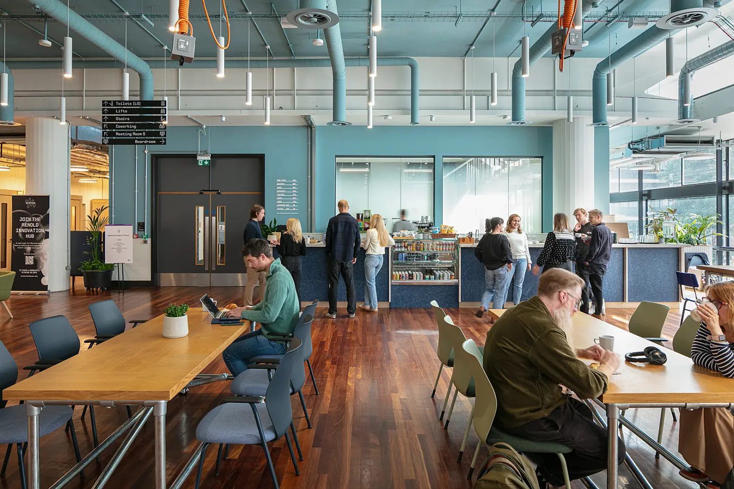 Busy workplace café and collaboration space with polished wood flooring, communal tables and a café counter serving drinks and refreshments. Several people are seated at tables working or talking, while others stand near the counter. The interior features a blue-painted exposed ceiling with visible ductwork, suspended lighting and hanging power outlets, alongside large windows that bring in natural light. Plants are positioned throughout the space, creating an informal environment for work, meetings and social interaction.