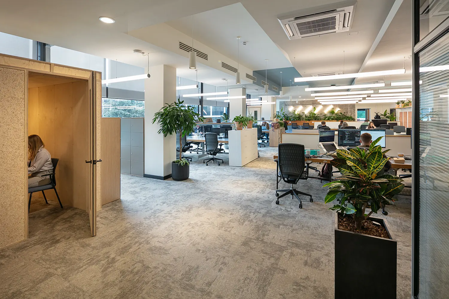 Open-plan office with a mix of workstations, collaborative areas and private focus booths. Employees are working at desks arranged throughout the space, which features grey carpet flooring, suspended linear lighting and large windows providing natural daylight. Integrated planting, storage units and timber finishes create distinct zones, while a wood-lined enclosed booth offers a quiet space for focused work or private calls. The interior combines contemporary workplace design with biophilic elements to create a bright and flexible working environment.