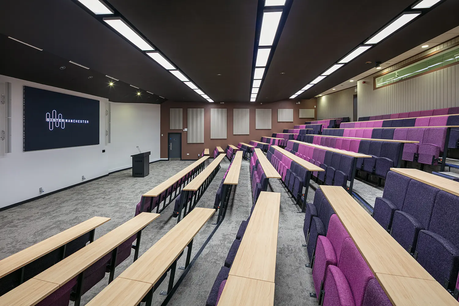 Tiered lecture theatre with rows of purple upholstered seating and light wood writing desks arranged in a stepped layout facing a presentation screen and lectern. The room features a dark ceiling with integrated linear lighting, acoustic wall panels and grey flooring. A large display screen at the front shows the Sister Manchster logo, while the overall design creates a modern teaching and presentation environment.