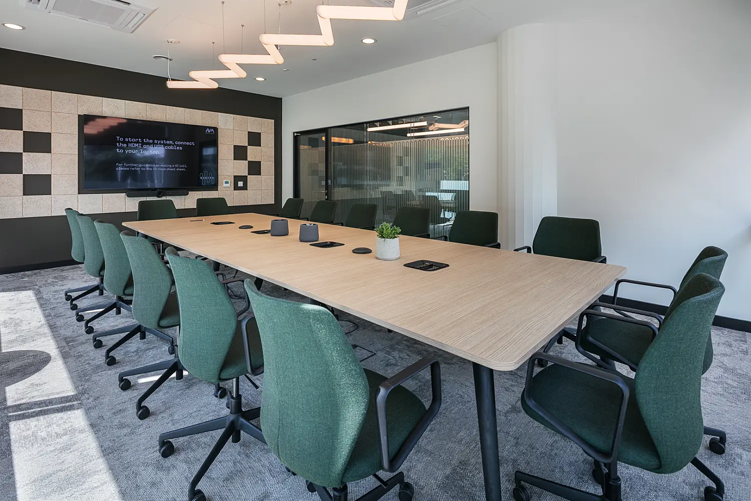 Contemporary meeting room with a large rectangular wood-finish conference table surrounded by green upholstered task chairs. Integrated power and connectivity modules are built into the tabletop, while a wall-mounted display screen and video conferencing equipment support hybrid meetings. The space features a patterned acoustic wall, glazed partition, grey carpet flooring and sculptural zigzag pendant lighting, creating a bright and modern collaborative environment.