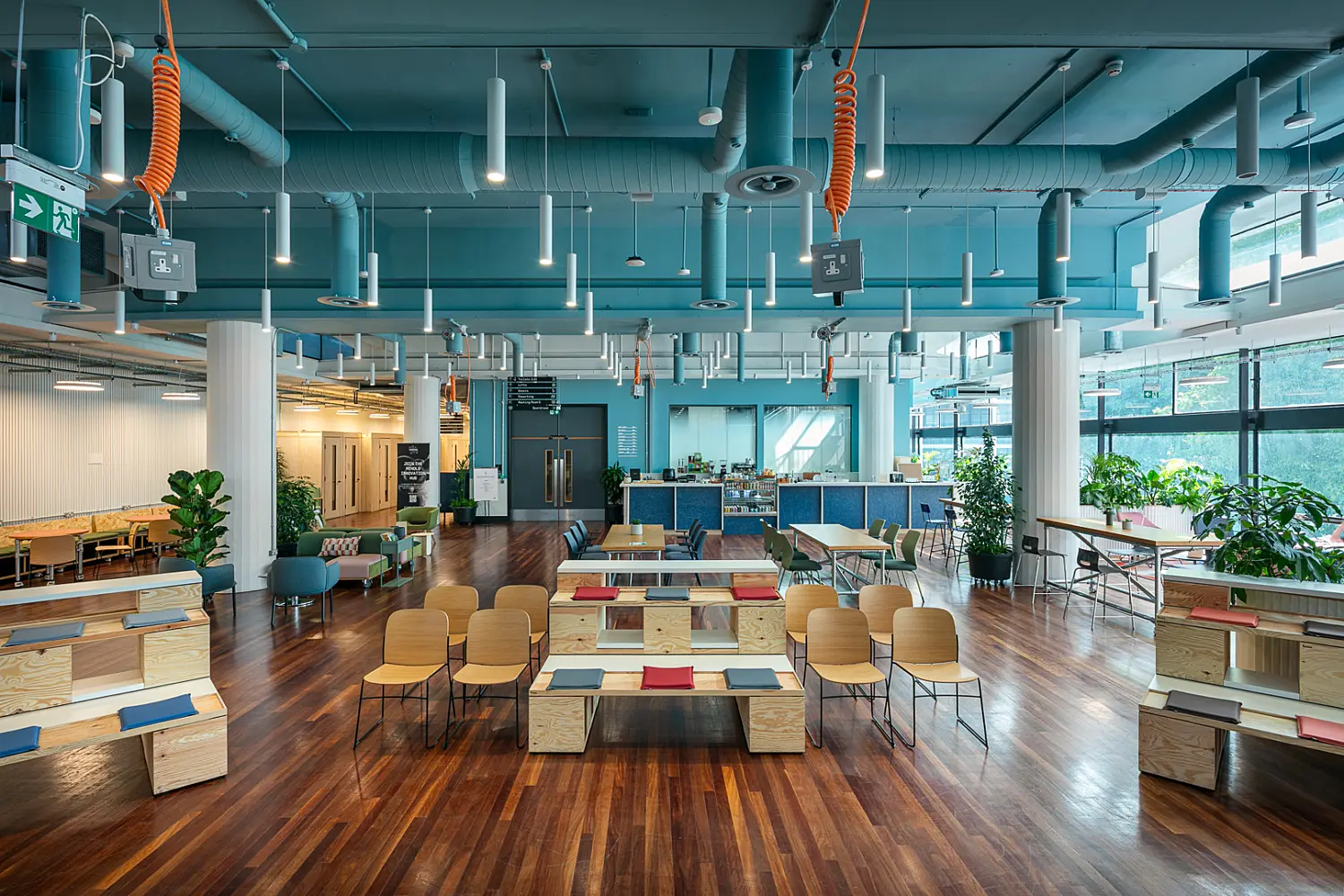 Open plan workplace lounge and collaboration space with polished wood flooring, tiered timber seating, tables and chairs arranged for informal meetings, and a café counter at the rear. The interior features a teal-painted exposed ceiling with visible ductwork, suspended lighting and hanging power outlets. Large windows along one side bring in natural light, while plants are placed throughout the space.