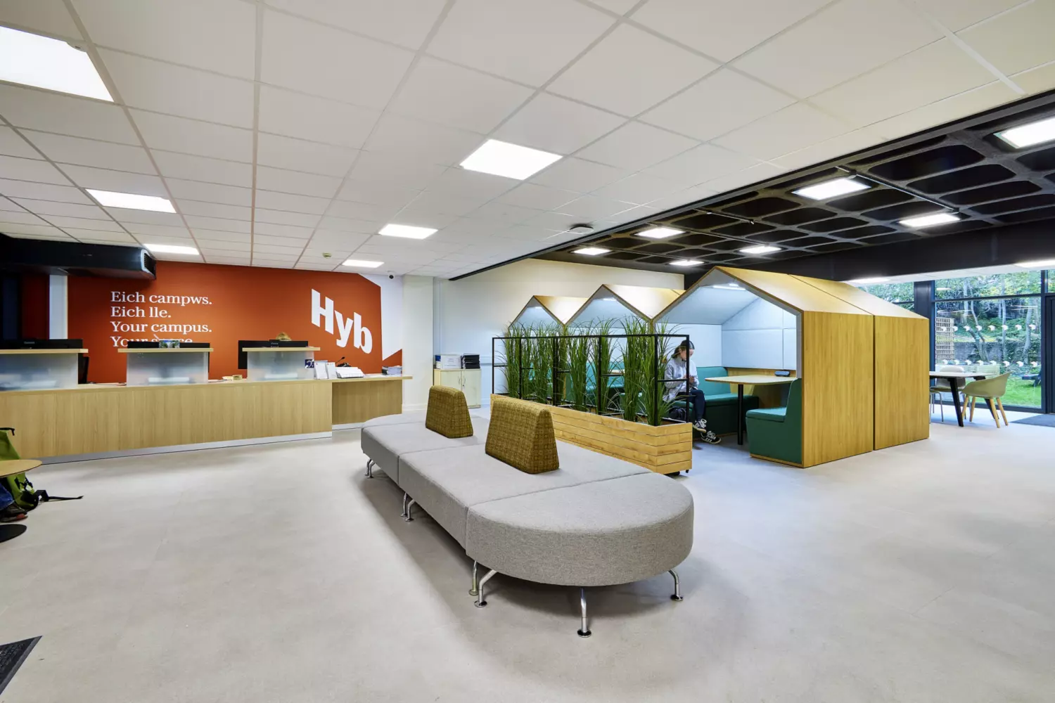 Reception and collaboration area with a modern fit out featuring a long wooden counter against a burnt orange accent wall with bold white text. The space includes semi-enclosed booths with pitched roof frames and green upholstered seating for private work. A central grey upholstered bench with two backrests provides informal seating. A planter with tall greenery adds a natural element and large windows bring in daylight. The design combines neutral flooring, suspended ceiling panels and clean lines to create a functional and welcoming environment.