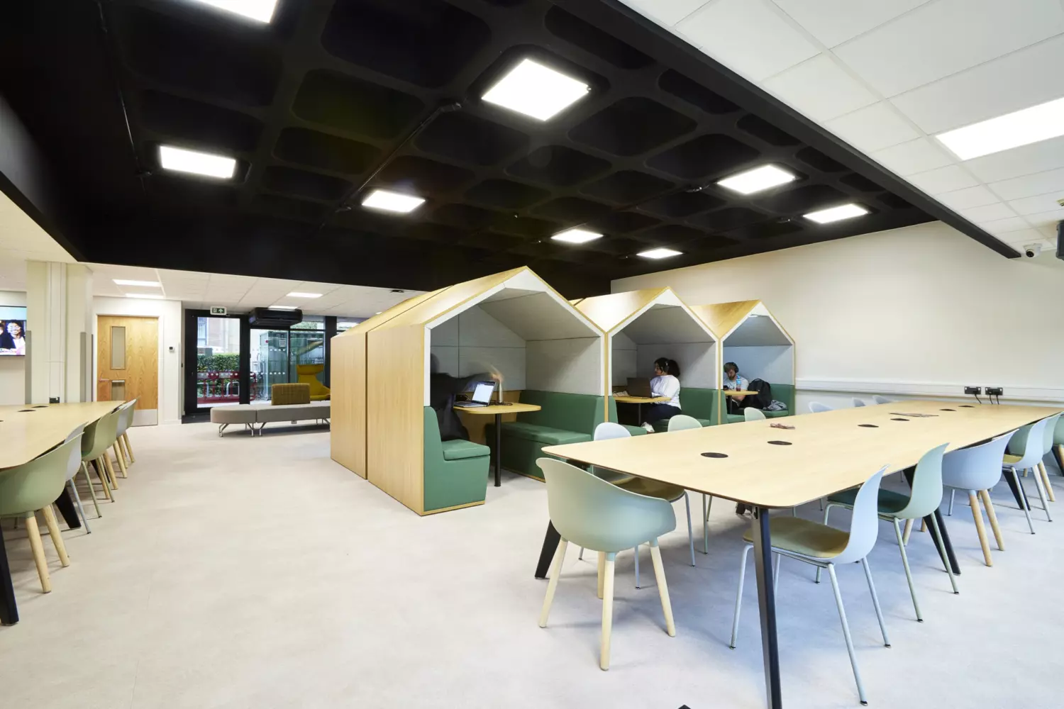 University collaboration area featuring long shared tables with integrated power points and mixed grey and green chairs. Three semi-enclosed booths with pitched roof frames and green upholstered seating provide private workspaces. The design includes neutral flooring, a black open ceiling grid with recessed lighting and clean white walls. Large windows at the far end allow natural light and the fit out combines functional furniture, simple lines and soft tones to create a practical and contemporary environment.