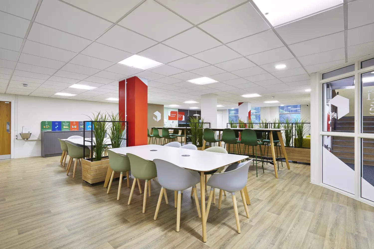 Open-plan university space with a modern fit out featuring white tables with integrated power points and mixed green and grey chairs. The design includes wood-effect flooring, suspended ceiling panels with recessed lighting and bright red accent columns. Planters with tall greenery create natural partitions between seating areas and high tables with bar stools provide additional collaborative zones. Large windows and glass partitions allow natural light and the overall layout combines clean lines and functional furniture for a contemporary and welcoming environment.