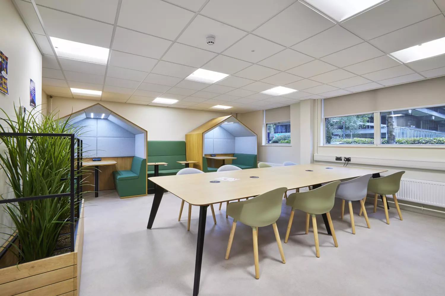 Modern university collaboration space featuring a large shared table with mixed grey and green chairs and integrated power points. Two semi-enclosed booths with pitched roof frames and green upholstered seating provide private work areas. The design includes neutral flooring, suspended ceiling panels with recessed lighting and wide windows allowing natural light. A planter with tall greenery adds a biophilic element and the fit out combines functional furniture, clean lines and soft tones to create a practical and inviting environment.