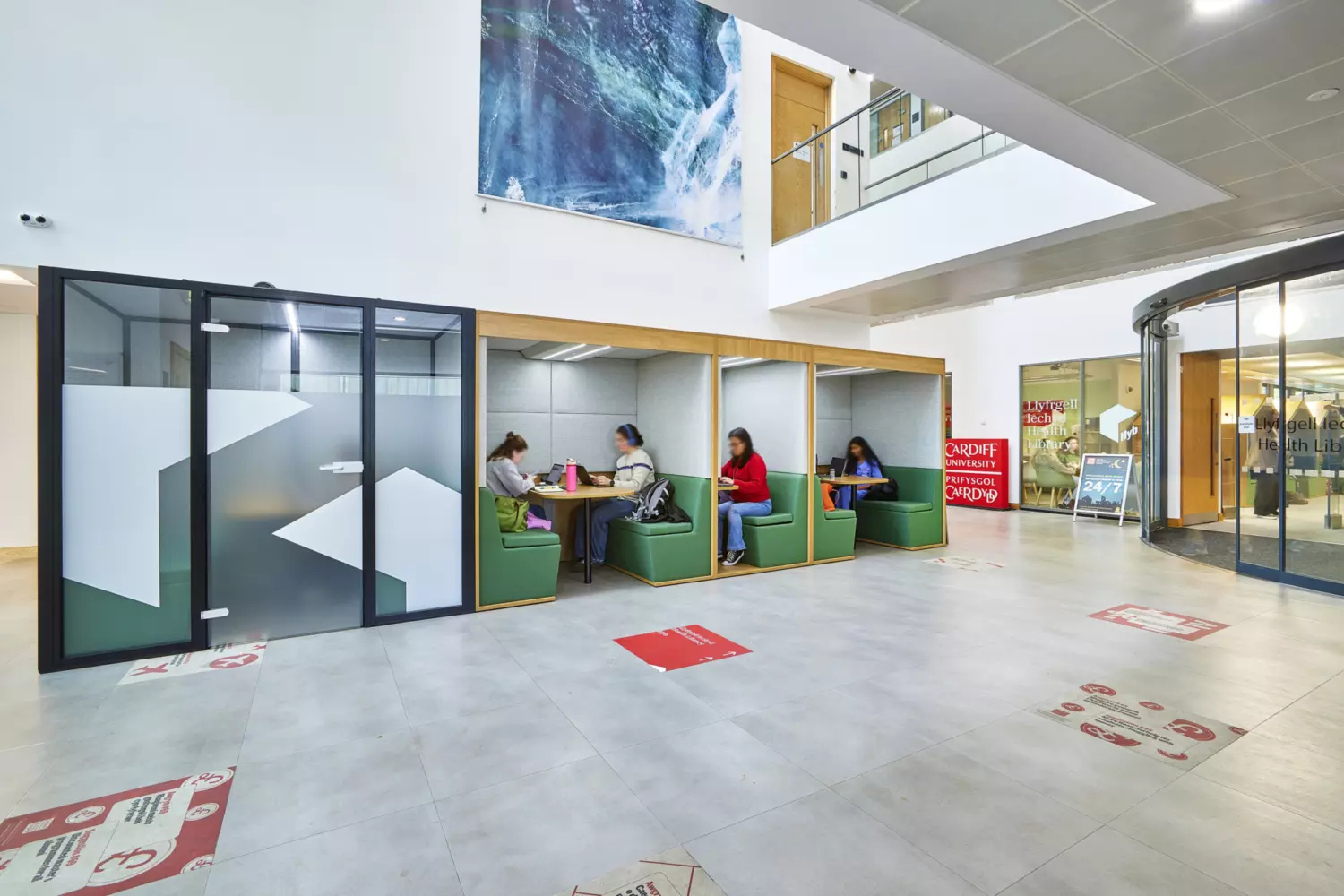 Contemporary university atrium featuring semi-enclosed booth seating for collaborative work. The design includes modular green upholstered booths with integrated tables set within a wooden frame structure. Frosted glass panels with geometric patterns provide partial privacy while maintaining an open feel. The space has light grey tiled flooring with directional graphics and a high ceiling that allows natural light. A large wall graphic adds visual interest above the booths and the fit out incorporates clean lines, neutral tones and functional furniture to create a modern and practical environment.