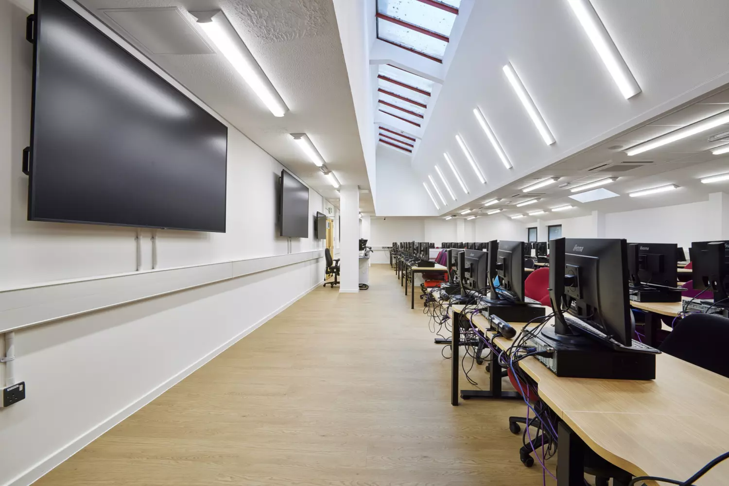 Computer lab with a modern fit out featuring rows of workstations with desktop monitors on light wood desks and black chairs. The space has white walls, wood-effect flooring and a high ceiling with integrated linear LED lighting. Large wall-mounted screens are positioned along one side and skylights allow natural light to enter. The design combines clean lines, neutral tones and functional furniture to create a bright and efficient working environment.
