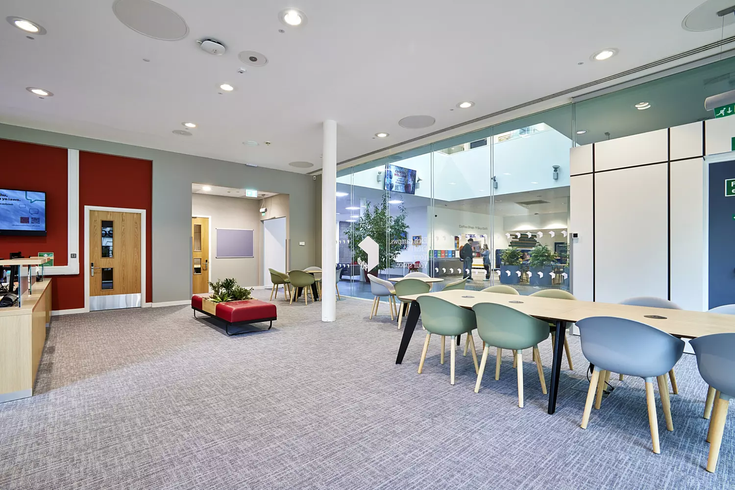 Modern open-plan university space featuring a mix of collaborative and breakout areas. The design includes a large glass partition wall creating transparency and natural light flow complemented by neutral grey carpeting and white ceilings with integrated circular acoustic panels. A long table with light green chairs provides a shared workspace while a red accent wall adds colour contrast near wooden doors. Soft seating in red and green tones is arranged around low planters for informal meetings. The fit out incorporates minimalist furniture, clean lines and biophilic elements with indoor plants creating a contemporary and welcoming environment.