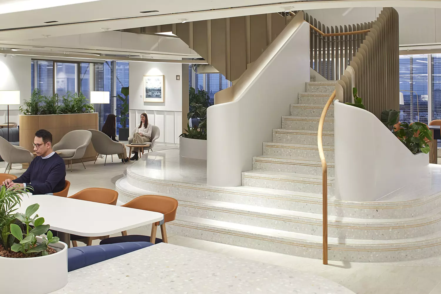 Well-lit interior space featuring a curved staircase that leads to an upper level. The area includes multiple seating arrangements with chairs and tables, some occupied by people. Large windows allow natural light to fill the room, while decorative plants add a touch of greenery. The overall design is open and inviting, combining functionality with visual appeal.