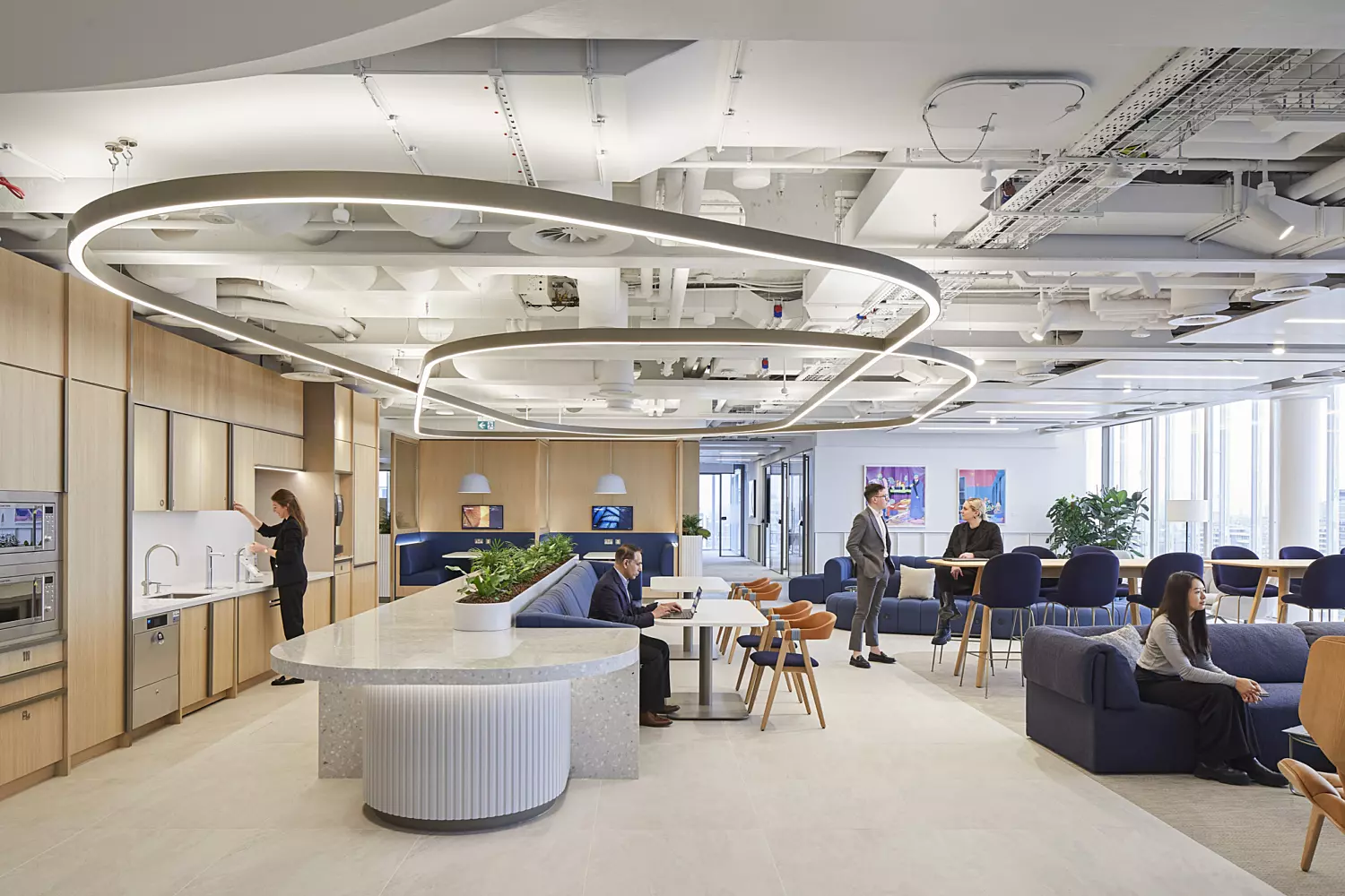 Office environment with a contemporary layout, featuring circular ceiling light fixtures, wooden panelling, and a variety of seating options including sofas and chairs. A central island with plants adds a natural element to the space. Several people are present, either working at tables or engaged in conversation. Large windows allow natural light to brighten the room, contributing to an open and inviting atmosphere.